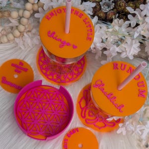 Op de afbeelding: Oranje en roze drinkgerei set met deksels, rietjes en onderzetters. De deksels hebben de tekst "Enjoy the little things" en "Run Dum Glucklich". De onderzetters hebben een bloemenpatroon en de tekst "Summer".