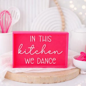 May include: A bright pink rectangular sign with the words "IN THIS kitchen WE DANCE" in white lettering. The sign is on a wooden surface, with kitchen utensils and dishes in the background.