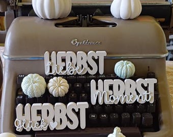 Puede incluir: Una máquina de escribir vintage con calabazas decorativas blancas y las palabras alemanas "Herbst Liebe Glück Moment". La máquina de escribir es de color marrón claro, y las calabazas son de varios tamaños y formas.