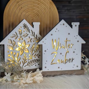 May include: Two white wooden house-shaped decorations with intricate cut-out designs. One features floral and leaf patterns, the other has the words "Winter Zauber" and star cutouts. Both are illuminated with warm light.