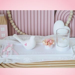Puede incluir: Una bandeja decorativa blanca con un pájaro de cerámica blanco con un corazón rosa, un pequeño arreglo floral y un portavelas de cristal. La bandeja está decorada con encaje y cinta rosa.