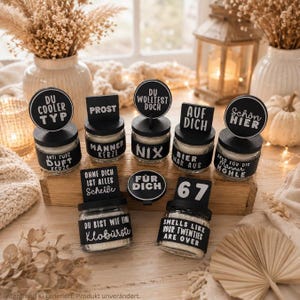 May include: A collection of seven small glass candles with black lids and labels. The labels feature white text in German and English, including phrases like 'Prost', 'Du cooler Typ', '67', and 'Smells like your twenties are over', arranged on a wooden block.