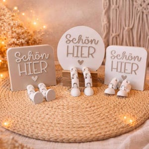 May include: Three decorative signs with the text "Schön HIER" and a heart symbol. The signs are white and beige, with attached legs and feet. The signs are arranged on a woven mat.