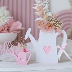 May include: Two white wooden watering cans with pink hearts. The larger watering can has dried flowers and a wooden sign that says "Endlich Zuhause". The smaller watering can is pink and has a small bouquet of dried flowers.