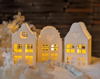 May include: Three white house-shaped candle holders with cut-out windows and heart details. Each structure features a warm yellow glow from within, creating a soft illumination. A string of wooden beads lies in front of the houses.