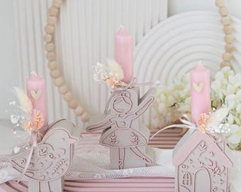 Puede incluir: Un conjunto decorativo con tres velas rosas, cada una en un soporte gris diferente: un pájaro, una bailarina y una casa. Las velas tienen detalles de corazón y están decoradas con flores secas y cintas, presentadas en una bandeja rosa.