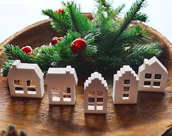 May include: Five small, white decorative house figurines with cut-out windows are arranged in a wooden bowl with green pine branches and red berries. The houses have varying roof designs, creating a festive holiday display.