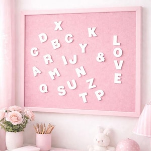 May include: A pink framed felt letter board with white letters spelling out words and letters. The word "LOVE" is spelled out on the right side. Other letters are scattered across the board. A vase of pink roses, pencils, and a bunny are in the foreground.