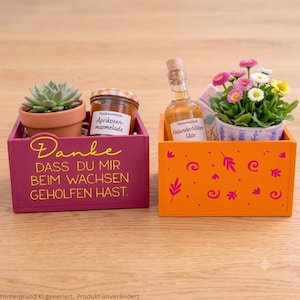 Puede incluir: Una caja de madera rosa oscuro con texto amarillo Danke dass du mir beim Wachsen geholfen hast que contiene una suculenta y mermelada de albaricoque, y una caja naranja con patrones florales rosas que contiene licor de saúco y margaritas.