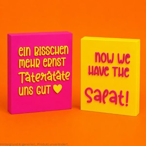 Pode incluir: Duas placas retangulares com letras em relevo. A placa da esquerda é rosa choque com texto amarelo "ein bisschen mehr ernst Täterätäte uns gut" e um coração amarelo. A placa da direita é amarela com texto rosa "now we HAVE THE Salat!"