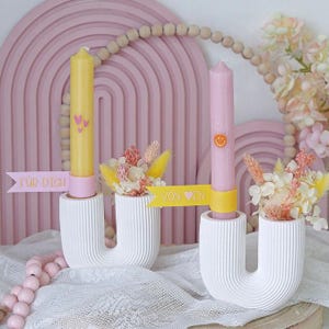 Könnte beinhalten: Zwei weiße, U-förmige Kerzenhalter mit gelben und rosa Kerzen. Die gelbe Kerze hat Herzmuster, und die rosa Kerze hat ein Smiley-Gesicht. Jeder Halter hat ein kleines Blumenarrangement und ein Banner mit den Worten "FÜR DICH" und "VON EN".
