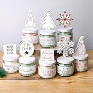 May include: A collection of glass candle jars with white lids, each topped with a unique decorative cutout. The jars feature labels with German text including Wichtel Gruß, Besinnlicher Scheiss, Frohe, Advent, and Anti Weihnacht Club.