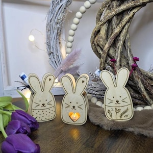 Puede incluir: Tres portavelas de madera con forma de conejito con detalles grabados. Los conejitos tienen orejas en forma de corazón y caras pintadas. Uno tiene un estampado de lunares, otro un recorte en forma de corazón y el tercero diseños florales. El fondo incluye una corona y tulipanes morados.