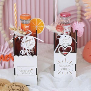 Könnte beinhalten: Zwei Flaschen Aperol Spritz Cocktails in dekorativen Haltern. Jede Flasche hat einen Strohhalm, Trockenblumen und ein Etikett. Ein Halter sagt "Summer Moments" und der andere "Summer Vibes". Eine Orangenscheibe befindet sich neben einer Flasche.