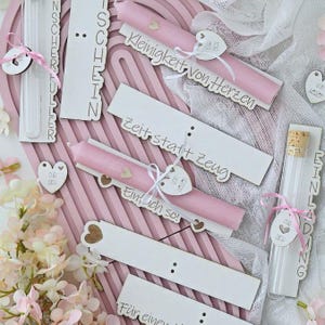 Puede incluir: Una colección de velas cónicas de color rosa claro y tubos de ensayo de vidrio con tapones de corcho, unidos a placas de madera blanca con texto en alemán cortado con láser como "Kleinigkeit von Herzen" y "Zeit statt Zeug," decorados con cintas rosas.