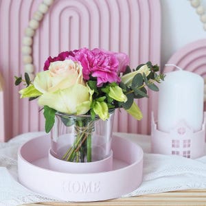 以下が含まれることがあります： 「HOME」と刻まれたピンクのセラミックトレイに、透明なガラスの花瓶が置かれ、ピンク、白、緑の花束が活けられています。