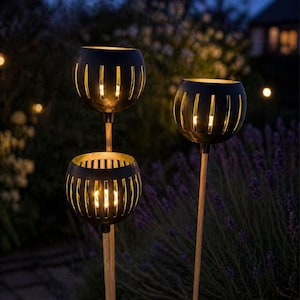 Peut inclure: Trois lanternes de jardin en métal noir avec un design sphérique et des découpes verticales, chacune avec une lumière chaude et brillante à l'intérieur. Les lanternes sont montées sur des poteaux en bois, sur un fond flou de feuillage et de lumière douce.