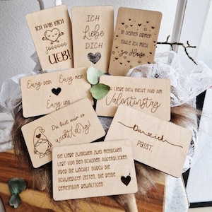 Op de afbeelding: Een verzameling houten kaarten met gegraveerde tekst en hartontwerpen. De kaarten bevatten zinnen als "Ik hou van je" en "Voor altijd wij". Sommige kaarten hebben illustraties van een hart en een kabouter. De kaarten zijn lichtbruin.