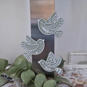 May include: Three decorative bird magnets in flight, each with a gray body and white outline. The birds feature detailed wing patterns and floral accents. The magnets are displayed on a metallic surface, with greenery in the foreground.