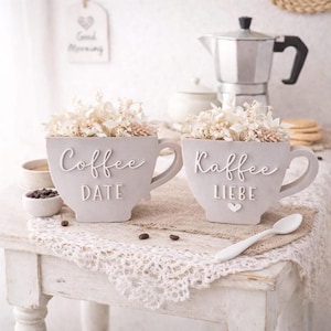Puede incluir: Dos macetas grises con forma de taza llenas de flores secas. Una dice "Coffee DATE" y la otra "Kaffee LIEBE". También se ven una cuchara blanca, granos de café y una cafetera, lo que sugiere una decoración temática de café.