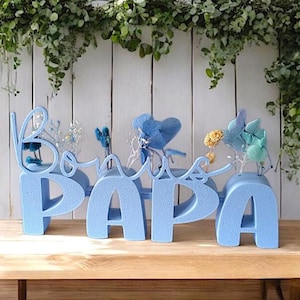 May include: Blue foam letters spelling "Bonjour Papa" with dried flowers in each letter. The letters are sitting on a wooden surface.