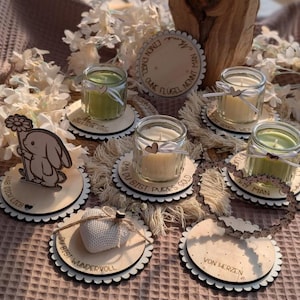 May include: A collection of small glass jar candles in various colors, resting on decorative wooden coasters. Some coasters feature laser-cut designs and text, with a heart-shaped accent on some candles. The overall theme is a rustic, handmade aesthetic.