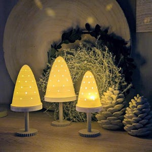 May include: Three cone-shaped lamps with white perforated surfaces emitting a warm yellow glow. Each lamp stands on a circular base and a central stem. Two textured pinecone-shaped ornaments are positioned to the right of the lamps.