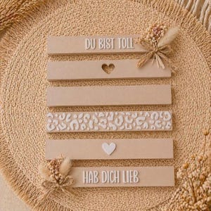 May include: Beige wooden decorative signs with white embossed text and designs. The signs include the phrases "DU BIST TOLLE" and "HAB DICH LIEB", along with heart and leopard print details. The signs are adorned with dried floral accents.