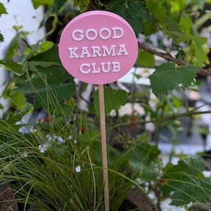Puede incluir: Un marcador de jardín rosa y circular con las palabras "GOOD KARMA CLUB" en letras blancas. El marcador está sobre un palo de madera, colocado en un jardín con hojas y hierba verdes.