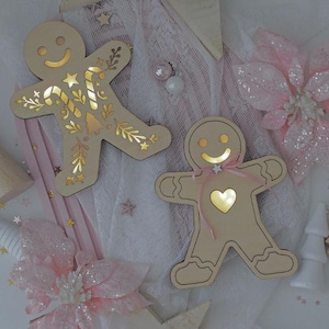 Puede incluir: Dos adornos de hombre de jengibre de madera con luces cálidas y brillantes. Uno tiene un diseño de bastón de caramelo y estrella, el otro un corazón. Ambos tienen una cara sonriente y están decorados con una cinta rosa y flores.