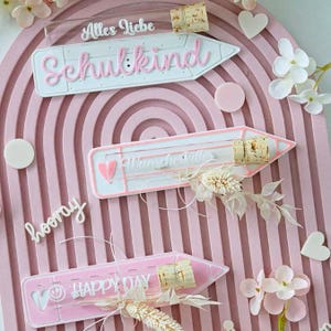 Puede incluir: Un tablero de exhibición arqueado de color rosa con tres placas en forma de lápiz y viales de vidrio con corcho. Las placas muestran el texto Alles Liebe Schulkind, Wunscherfüller y HAPPY DAY, decoradas con flores blancas y corazones.