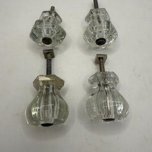 May include: Four clear glass cabinet knobs with a faceted, multi-sided top section and a rounded, ribbed base. Each knob features a central metal bolt with a square metal plate at the base of the screw, providing a secure mounting mechanism.