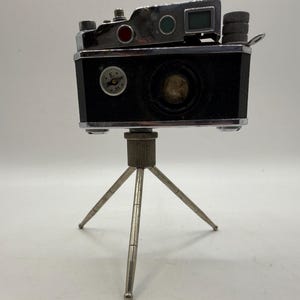 May include: A vintage black camera with silver trim, mounted on a small silver tripod. The camera has a lens, dials, and a viewfinder. It features red and green accents and a small compass-like emblem. The tripod has three adjustable legs.