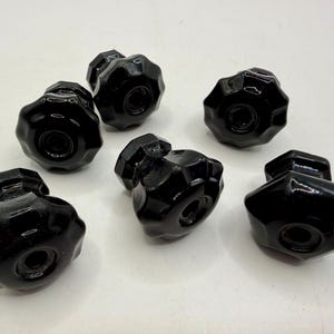 May include: A collection of seven glossy black knobs with a starburst design. Each knob has a central hole and a faceted, rounded shape. The knobs are arranged on a white surface, showcasing their reflective quality.