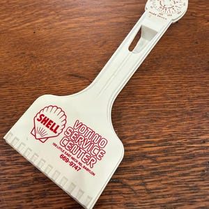 May include: A white plastic ice scraper featuring a red Shell logo and the text Votino Service Center, 199 Little East Neck Rd, Babylon, 669-9747. The handle includes a circular gauge for tracking miles driven and gallons used.