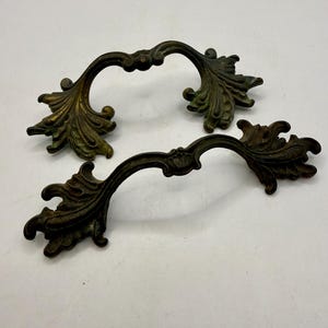 May include: Two antique bronze drawer pulls with a curved design. Each pull features ornate leaf and scroll details, with a slightly aged, weathered appearance. The pulls are approximately 5 inches wide.