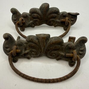 May include: Two matching dark brown metal drawer pulls with a weathered, rusted finish. Each pull features an ornate, symmetrical backplate with scrollwork details and a curved, textured drop handle attached by two metal loops and threaded bolts.