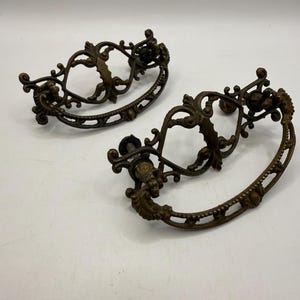 May include: Two matching antique-style drawer pulls made of dark bronze-toned metal. Each handle features an ornate, openwork design with intricate scrollwork and a curved, textured grip. The hardware includes two mounting posts on each piece.