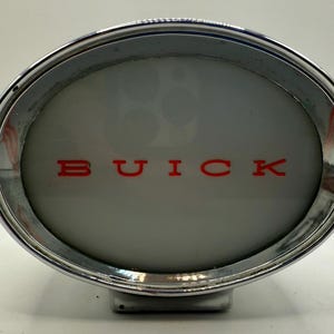 May include: Oval-shaped chrome car emblem with the word "BUICK" in red lettering. The emblem has a silver-gray background and a shiny chrome border. The emblem is likely from a vintage Buick vehicle.