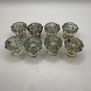 May include: A set of eight clear glass knobs arranged in two rows of four. Each knob features a multi-faceted, star-like design with a circular center indentation and a short, tapered base. The glass shows slight signs of age and minor discoloration.