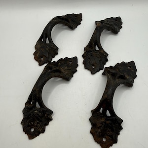May include: A set of four dark brown metal drawer pulls, each featuring a curved handle design with detailed leaf-shaped mounting plates at both ends. Each plate includes a single circular screw hole for attachment to a surface.