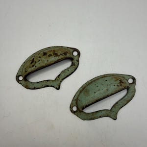 May include: Two metal drawer pulls with a weathered, light blue-green finish and visible rust spots. Each handle features a curved, semi-circular grip area and two screw holes on the flat mounting flanges for attachment to furniture surfaces.
