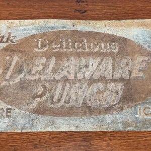 May include: Vintage Delaware Punch sign with faded text. The sign reads "Drink Delicious Delaware Punch" in a central oval, with "Sold Here" and "Ice Cold" at the bottom. The sign has a weathered, aged appearance.