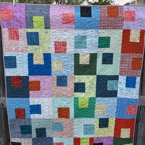 May include: A colorful patchwork quilt with a geometric design. The quilt features squares in various colors, including pink, blue, green, and orange. Each square has a smaller square in a contrasting color. The quilt is hanging on a wooden fence.