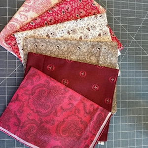May include: A collection of folded fabric squares in various shades of red, cream, and gray. The fabrics feature paisley and floral patterns. The fabrics are arranged on a gray cutting mat.