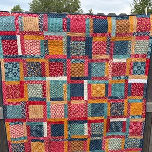May include: A colorful patchwork quilt with a grid pattern. The quilt features squares of various floral and patterned fabrics in shades of red, blue, yellow, and green. The quilt is hanging on a wooden fence.