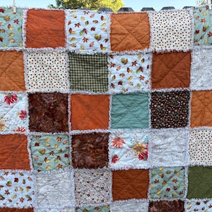 May include: A patchwork rag quilt featuring a grid of square fabric blocks in shades of orange, sage green, brown, and white. The quilt displays various patterns including autumn leaves, floral prints, polka dots, and solid colors with frayed seams.