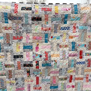 May include: A handmade patchwork quilt in a grid pattern. The quilt uses a variety of fabrics in beige, grey, teal, red, pink, and black. The rectangular blocks create a textured and colourful textile, perfect for a bed or sofa.
