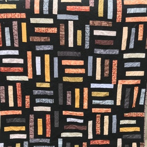 May include: A handmade quilt with a black background and a pattern of rectangular blocks in various colors. The blocks include shades of orange, brown, gray, and yellow. The quilt is displayed against a wooden fence.