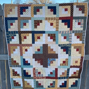 May include: A patchwork quilt with a log cabin pattern in shades of brown, blue, red, and cream. The quilt is made up of squares of different fabrics, each with a unique pattern.
